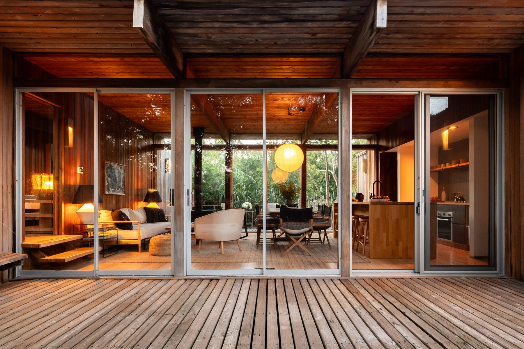 The house seen from the deck at dusk, sliding glass doors open onto the lit living and dining rooms