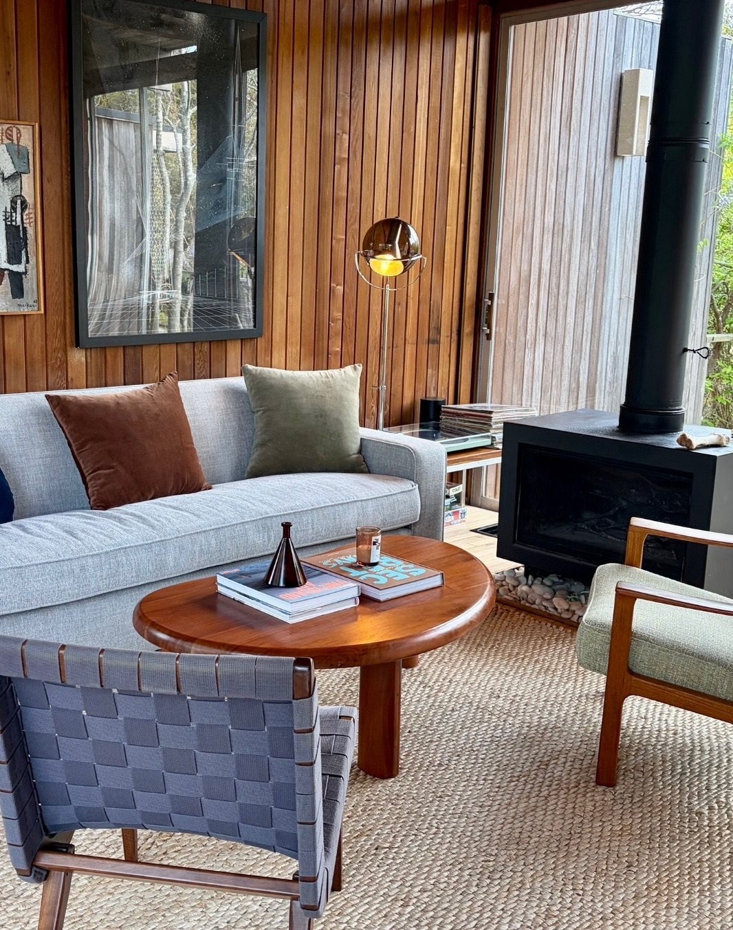 Living room with a grey sofa, round teak table and a black wood stove against cedar walls